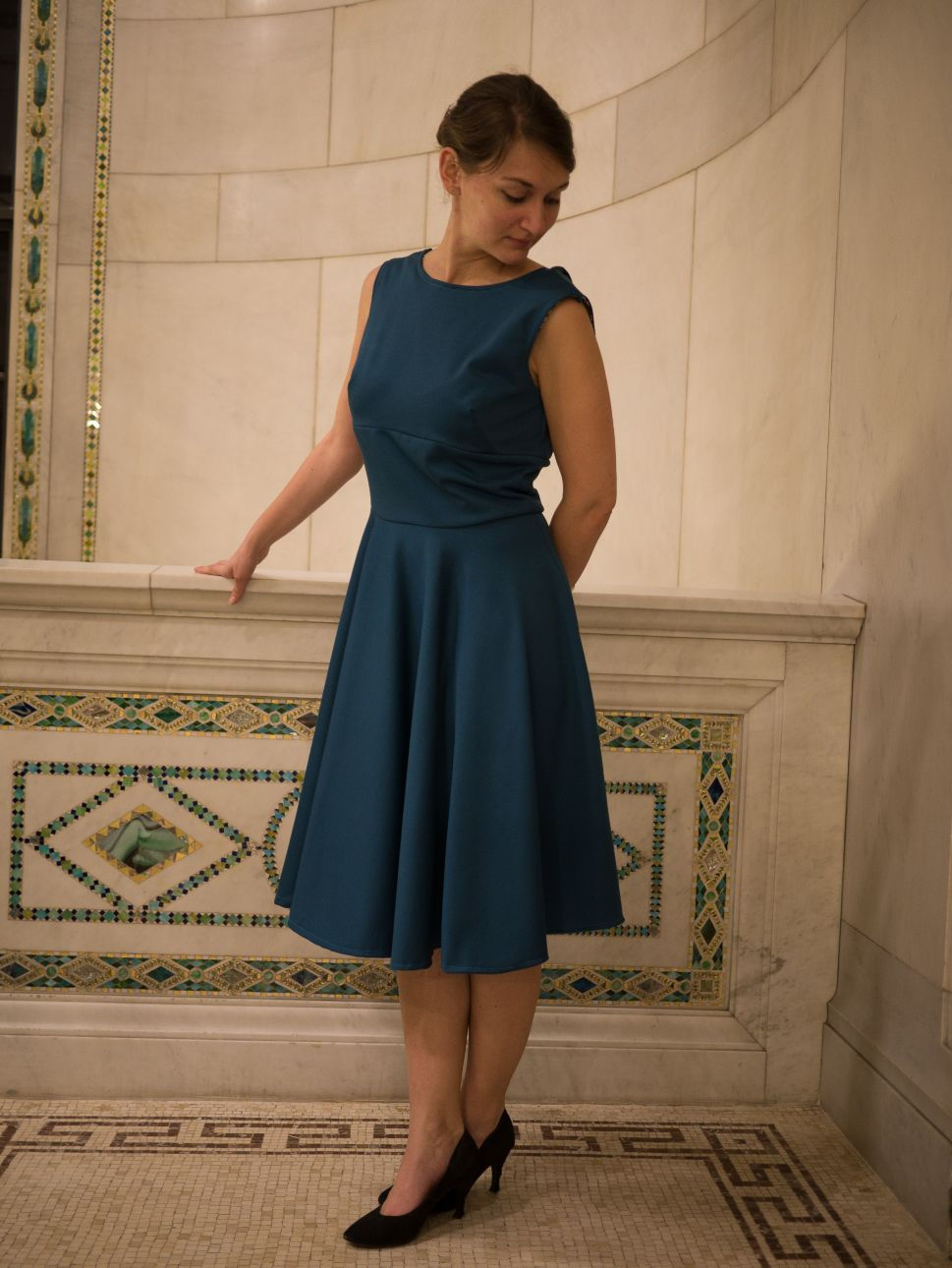 Une femme portant une robe col bateau et jupe circulaire, bleue, pose sur un balcon en mosaique.