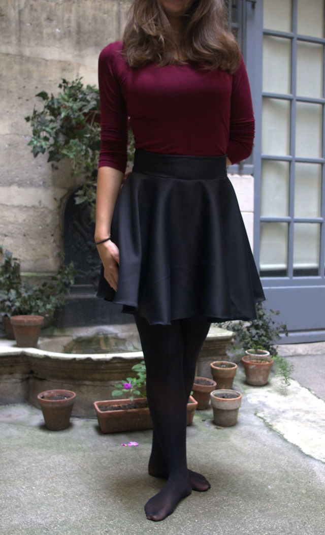 Une femme portant un tee-shirt bordeaux et une jupe patineuse noire, pose devant une fontaine.