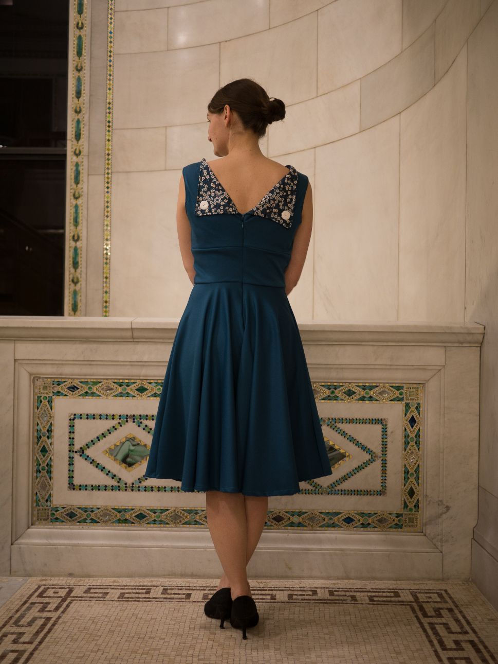 Une femme portant une robe col bateau et jupe circulaire, bleue, pose sur un balcon en mosaique.
