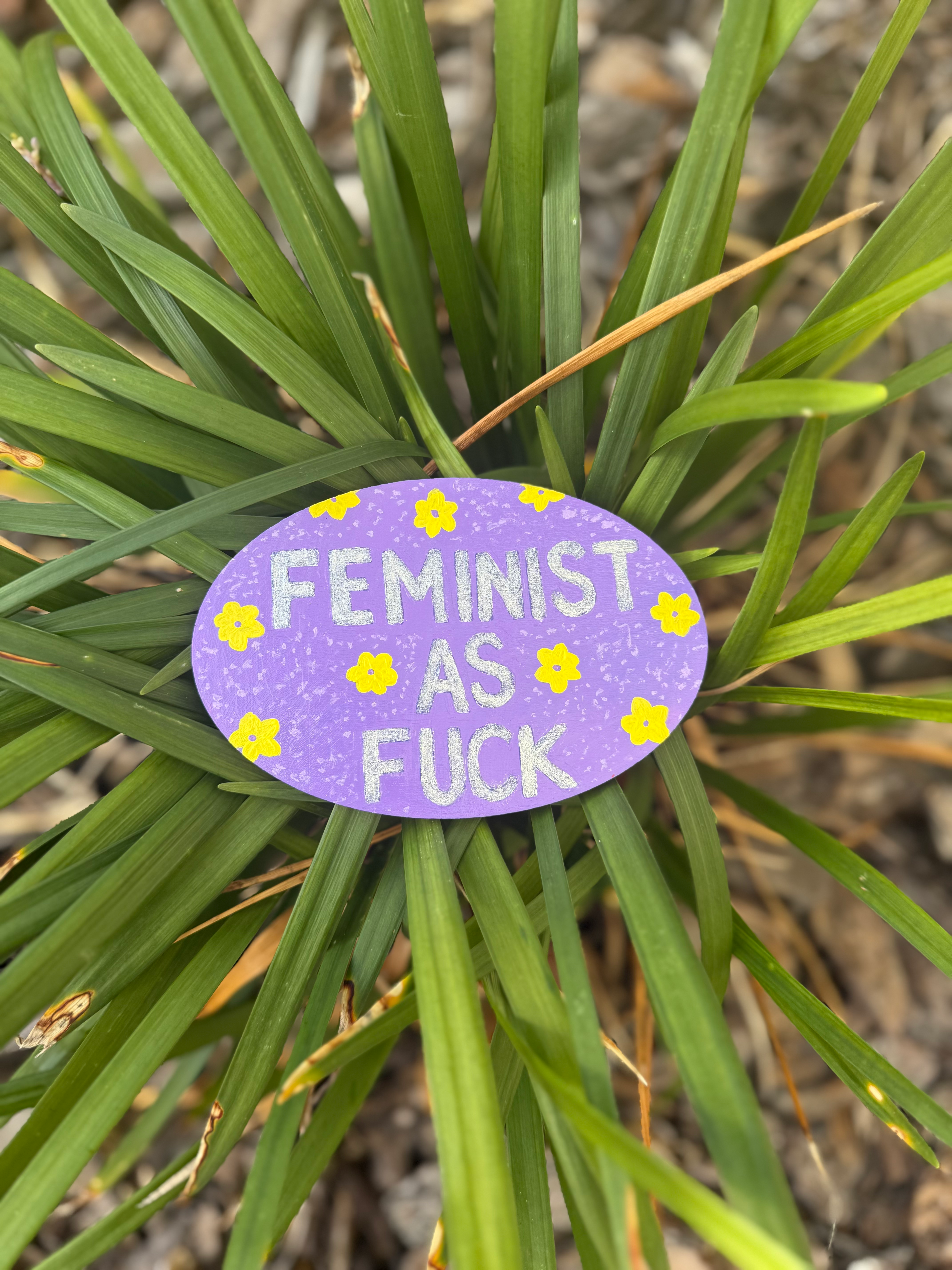 purple background with yellow flowers and glitter text that says "Feminist As Fuck"