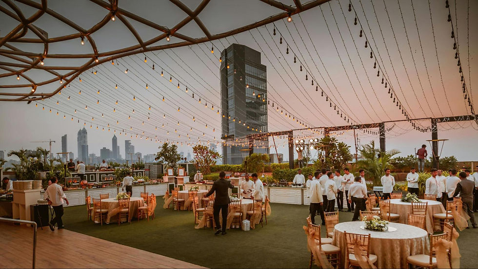 Rooftop event at sunset with string lights. People in white attire gather, tables set with flowers, buildings in the background. Festive mood.
