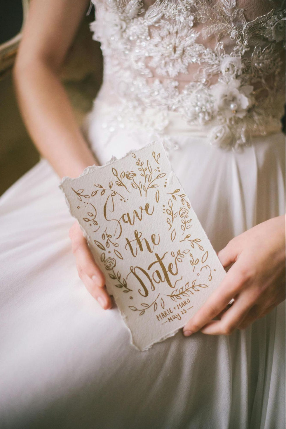 Bride in a lace dress holds a "Save the Date" card with golden floral design. Romantic and elegant setting.