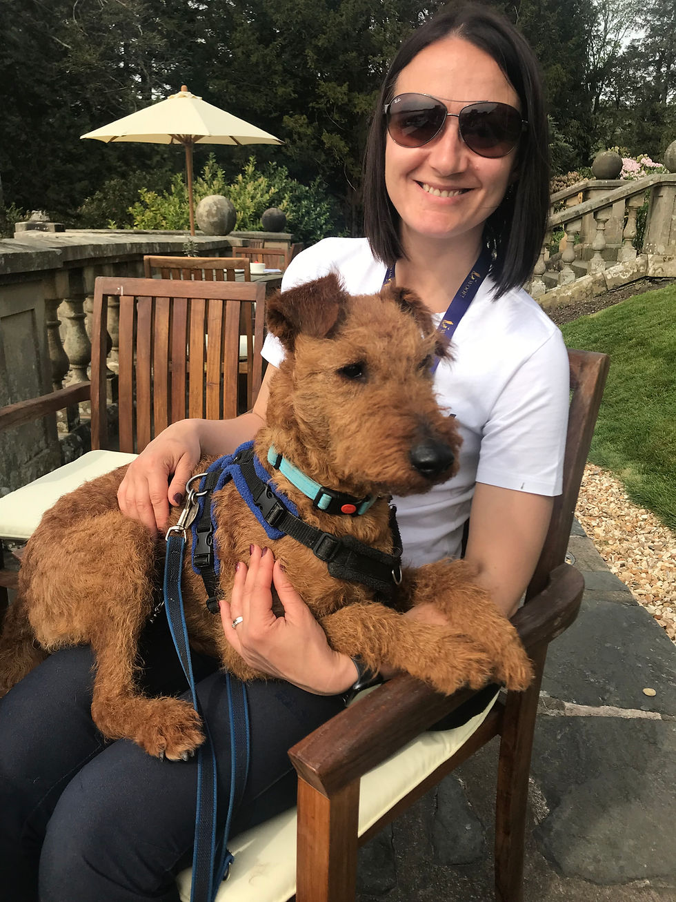 Tash in sunglasses smiling, holding Monty, a wire-haired tan Irish Terrier in a harness, on her lap at an outdoor table