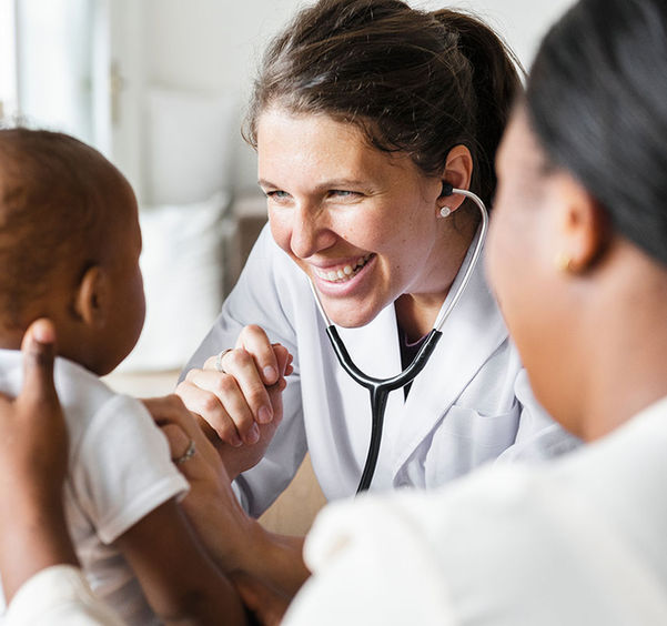 Female doctor smiling, checking heartbeat of infant patient with mother standing by.