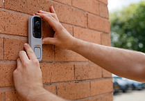 A person installs a sleek, modern video doorbell on a red brick wall