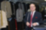 Man in pinstripe suit with measuring tape in tailor shop, surrounded by suits and ties on racks. He smiles, creating a warm, professional mood. Located in North End Halifax.