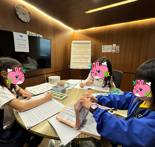 Three children sit around a table in a classroom, working on worksheets and using colored pencils, with a whiteboard and cloc