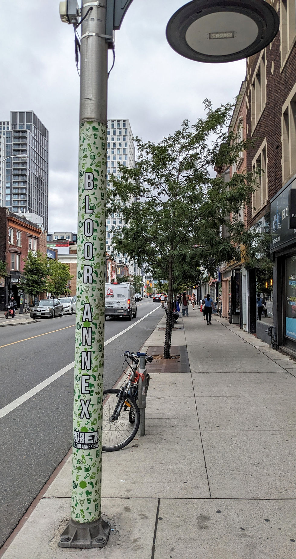 Street scene with a pole reading "BLOOR ANNEX," bike parked nearby, tree-lined sidewalk, buildings, and passing traffic on a cloudy day.