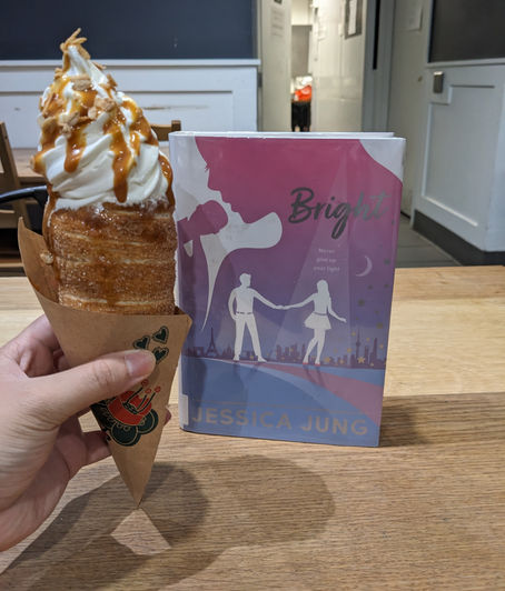 Hand holding a churro ice cream cone with caramel drizzle next to the book "Bright" by Jessica Jung on a wooden table in a cafe.