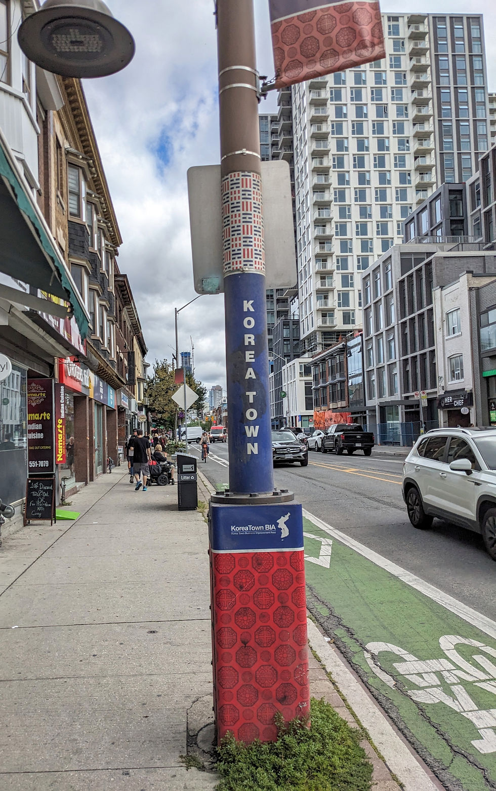 Street in Koreatown with a blue and red decorated pole. Buildings and cars line the street. Text reads "KOREA TOWN" and "KoreaTown BIA."