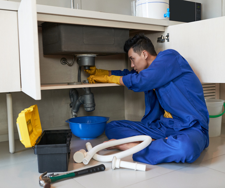A man wring blue overalls and yellow gloves working under a kitchen si.