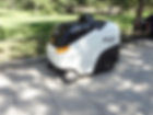 white and black robot on 4 wheels with 2 sweepers at the front, moving on a sidewalk