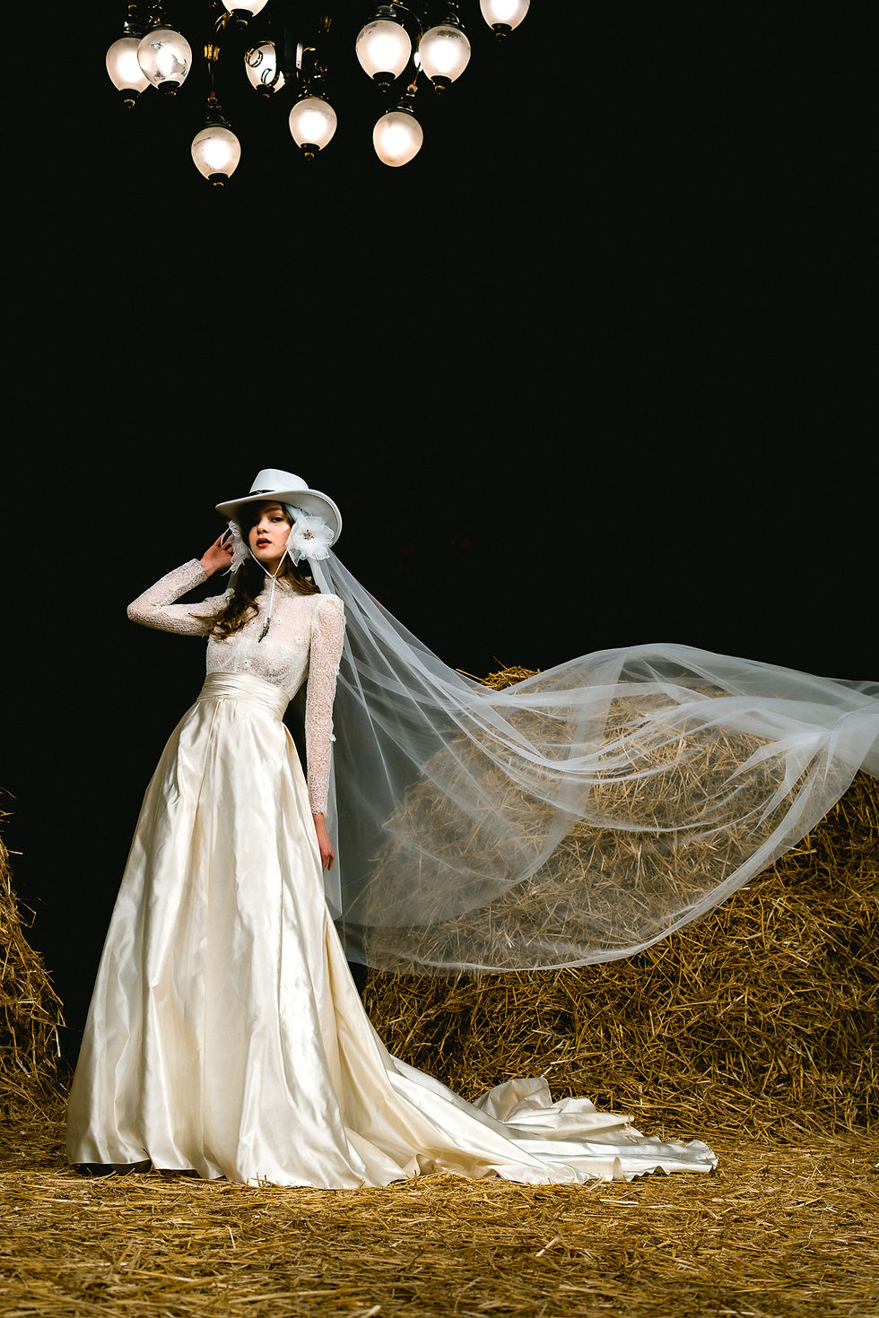 A model exudes elegance and rustic charm in the Aadnevik Autumn/Winter 2025 "Western Star" collection, featuring a flowing satin gown paired with a stylish cowboy hat and sheer veil, set against a backdrop of haystacks under vintage lighting.