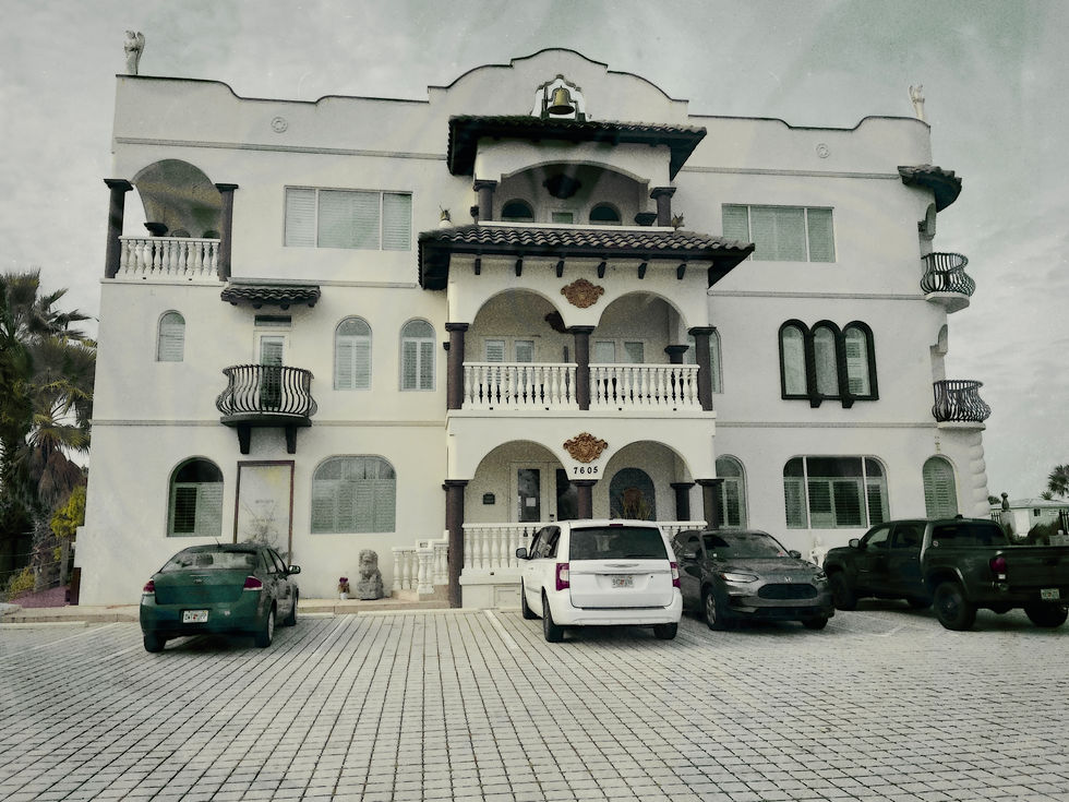 Large white building with ornate balconies and statues. Several parked cars in the cobblestone lot. Cloudy sky. Address: 7605.