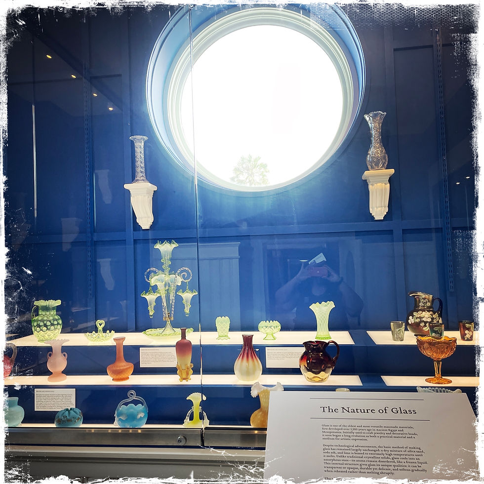 Glass display with vases and pitchers on shelves, set against a blue background. A round window and a plaque titled "The Nature of Glass."