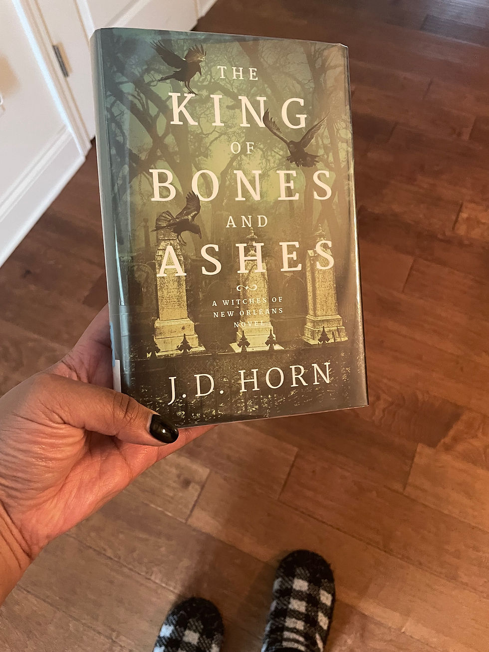 A hand holds the book "The King of Bones and Ashes" by J.D. Horn against a wood floor. The cover shows a dark, eerie forest scene.