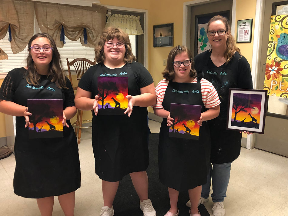 3 young women with Down syndrome stand with Cristin DeCamville, all wearing DeCamville Arts aprons and holding their paintings of giraffes in Africa at sunset.
