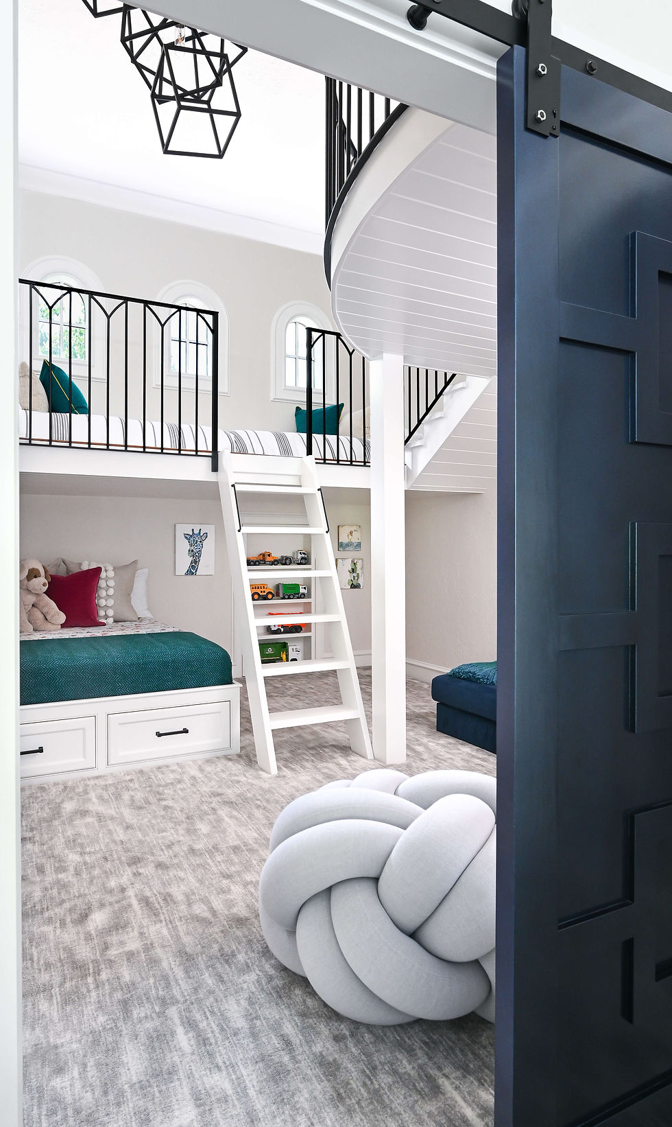 Bright kids’ loft bedroom with bunk beds, ladder, toys, teal and white bedding, black railings, and a large knot floor cushion.