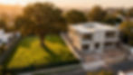 Vista aérea de una residencia moderna de concreto en construcción junto a un terreno verde con un gran roble centenario. La casa muestra una estructura minimalista con andamios, ubicada en una zona urbana residencial durante el atardecer.