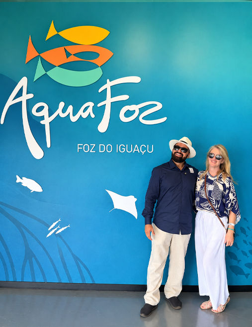 Marta e Anderson na entrada do AquaFoz, em Foz do Iguaçu, em frente ao painel azul com o logotipo do aquário, durante visita para a Revista EDVD.