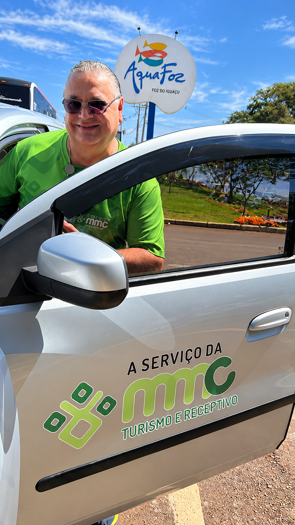 Representante da MMC Turismo ao lado de um veículo da empresa, com a identificação do AquaFoz ao fundo, em Foz do Iguaçu.