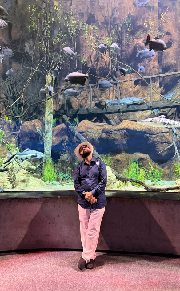 Anderson pousando para foto em frente ao tanque com peixes, troncos e vegetação aquática durante a visita ao AquaFoz, em Foz do Iguaçu.