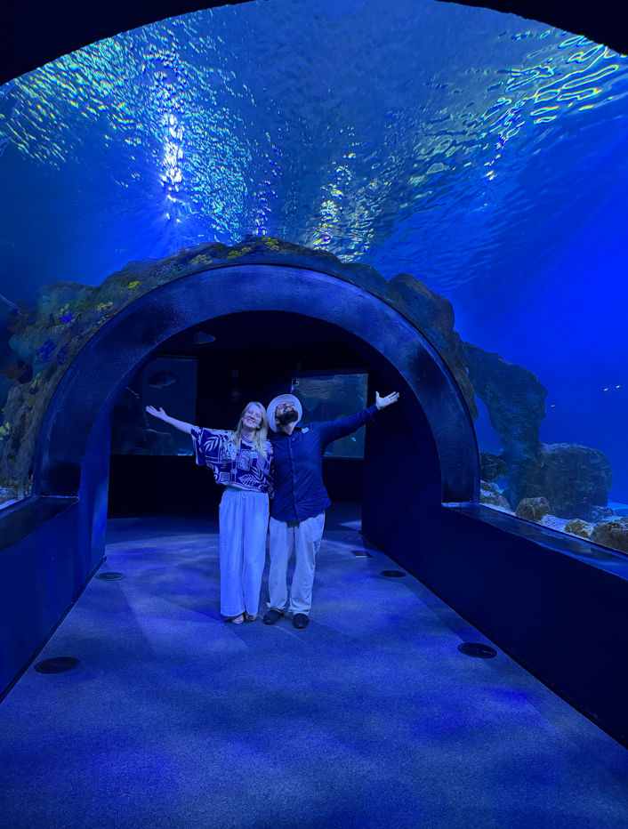 Marta e Anderson de braços abertos no túnel subaquático do AquaFoz, cercados pela iluminação azul e pelos grandes tanques do aquário.
