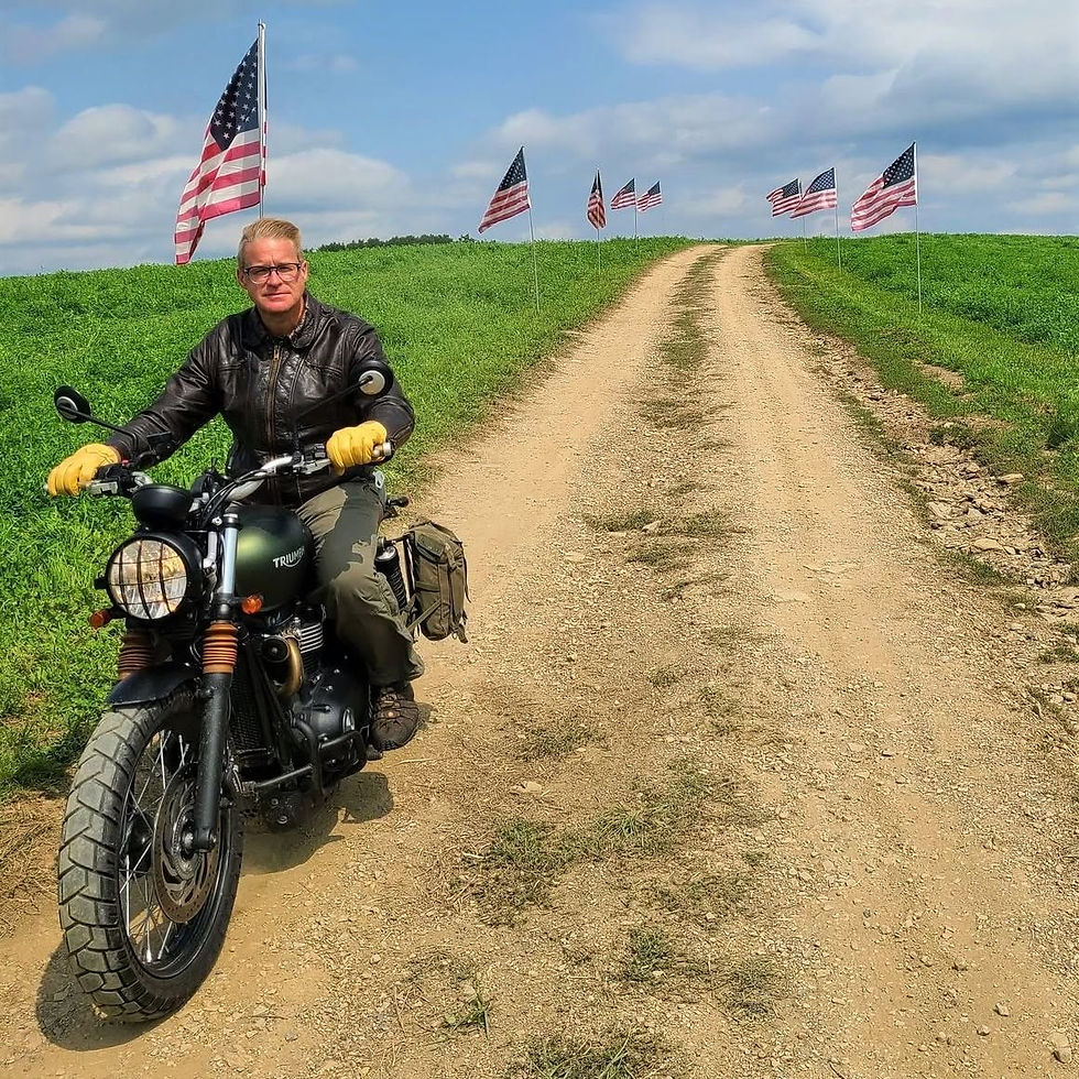 Scot Loveland riding his motorcycle down a dirt path lined with American Flags on a hill in a field.