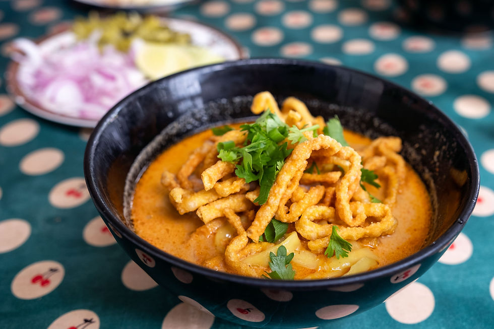 Un bol noir contenant du Khao Soi, un curry thaï du Nord au poulet, garni de nouilles croustillantes et de coriandre fraîche. En arrière-plan, une assiette d’accompagnements avec oignons rouges émincés, citron vert et légumes marinés, sur une table à motif thaïlandais.