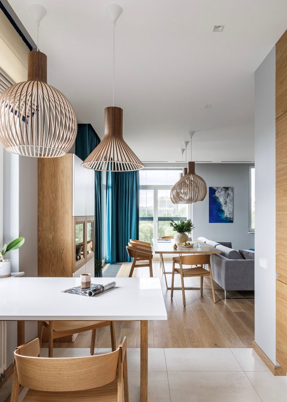 Stylowy salon z eleganckimi drewnianymi akcentami, szarym narożnikiem i niebieskim krzesłem, z dużymi, geometrycznymi lampami.