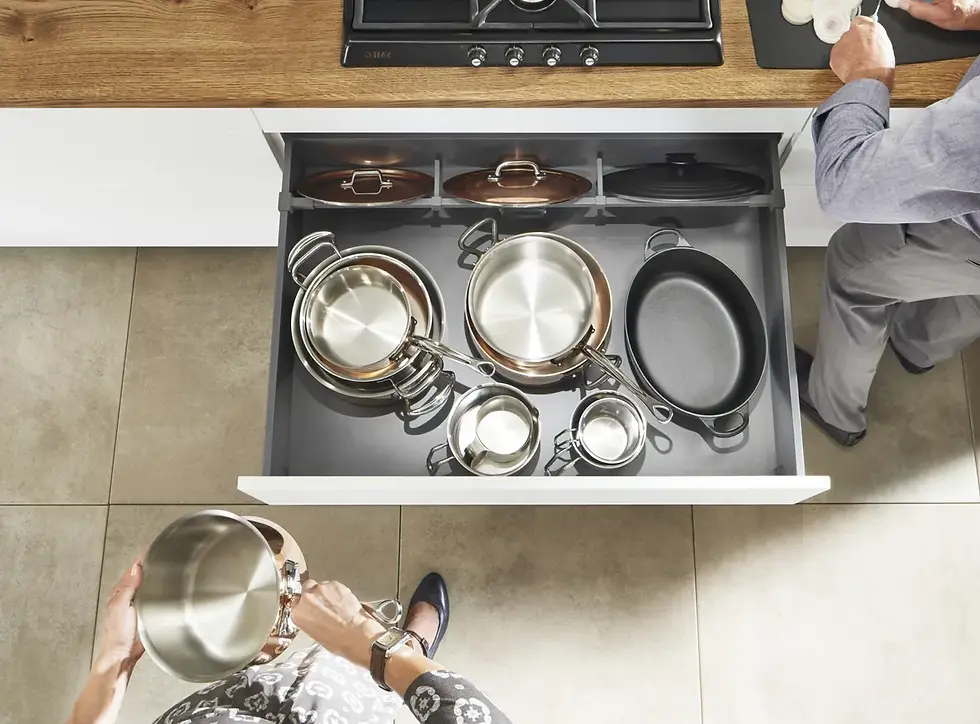 Kuchnia z szufladą pełną metalowych garnków, osoba trzyma garnek. Obok osoba kroi warzywa na desce. Jasne płytki i drewniany blat.