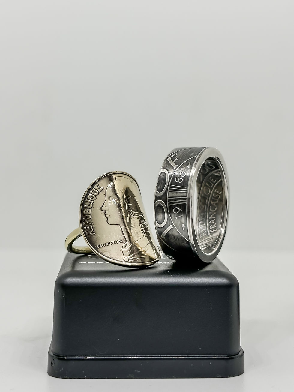 Bague et anneau en argent et or, faits de pièces françaises, posés sur un socle noir. Motifs et inscriptions visibles, fond blanc.