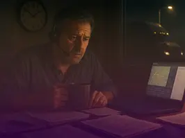 Stressed fleet manager working late at night with a headset and coffee, reviewing paperwork next to a laptop showing a truck tracking map.