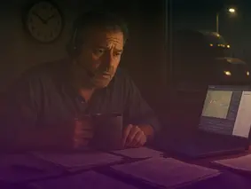 Stressed fleet manager working late at night with a headset and coffee, reviewing paperwork next to a laptop showing a truck tracking map.