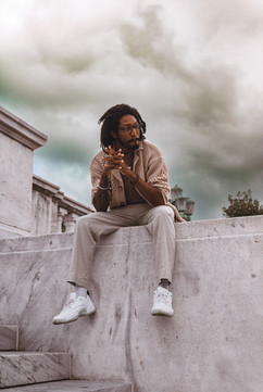 A man seated on a stone ledge, loooking off into the distance while a storm rolls in behind him.