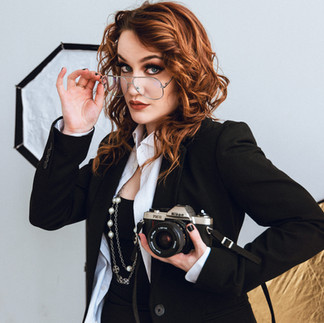 Woman holding a camera, tilting her glasses forward, in front of assorted photography equipment