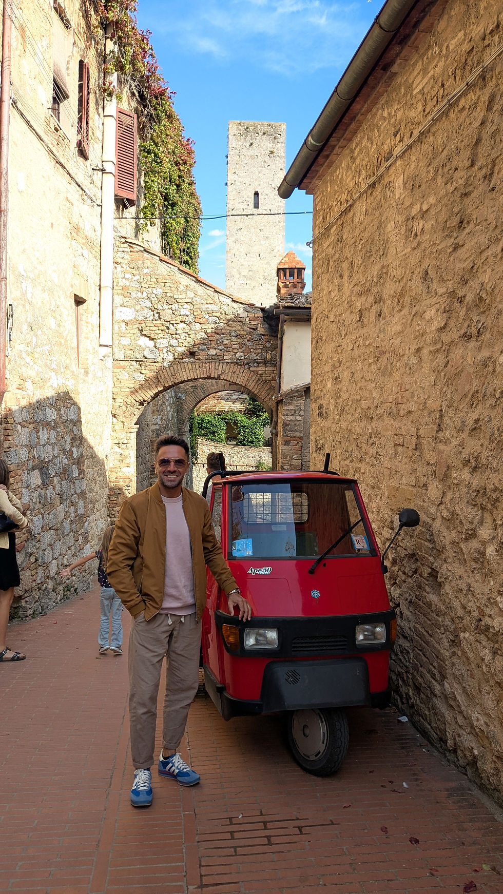 Walter DiLello standing beside a red Ape vehicle in a narrow stone alleyway in an Italian village, capturing authentic daily life and local culture in Italy.