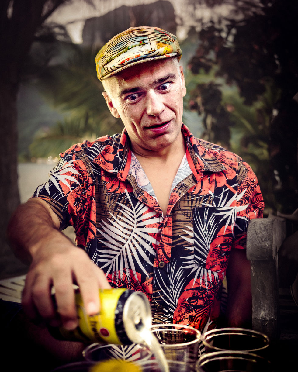 Un homme en chemise hawaïenne et casquette verse une canette de boisson dans des verres avec une expression drôle et expressive.