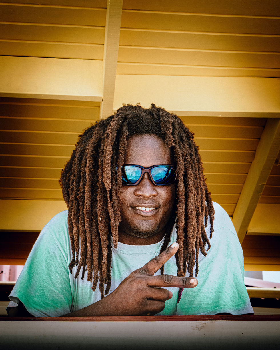 Homme aux dreadlocks portant des lunettes de soleil miroir et faisant un signe peace sous une structure en bois à Saint-Martin, portrait lifestyle tropical et urbain.