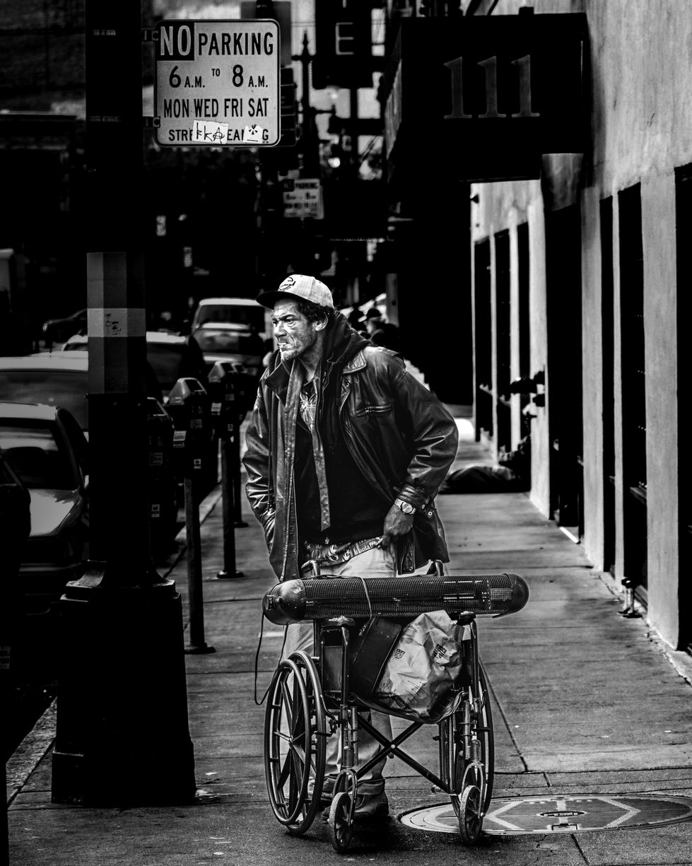 Photographie de rue contrastée en noir et blanc à San Francisco, montrant un homme de profil portant une casquette, une veste en cuir et un sweat à capuche. Il se tient sur le trottoir à côté d'un fauteuil roulant chargé d'effets personnels. En arrière-plan, on aperçoit des panneaux de signalisation urbaine, dont un panneau "No Parking", et la perspective d'une rue de la ville.