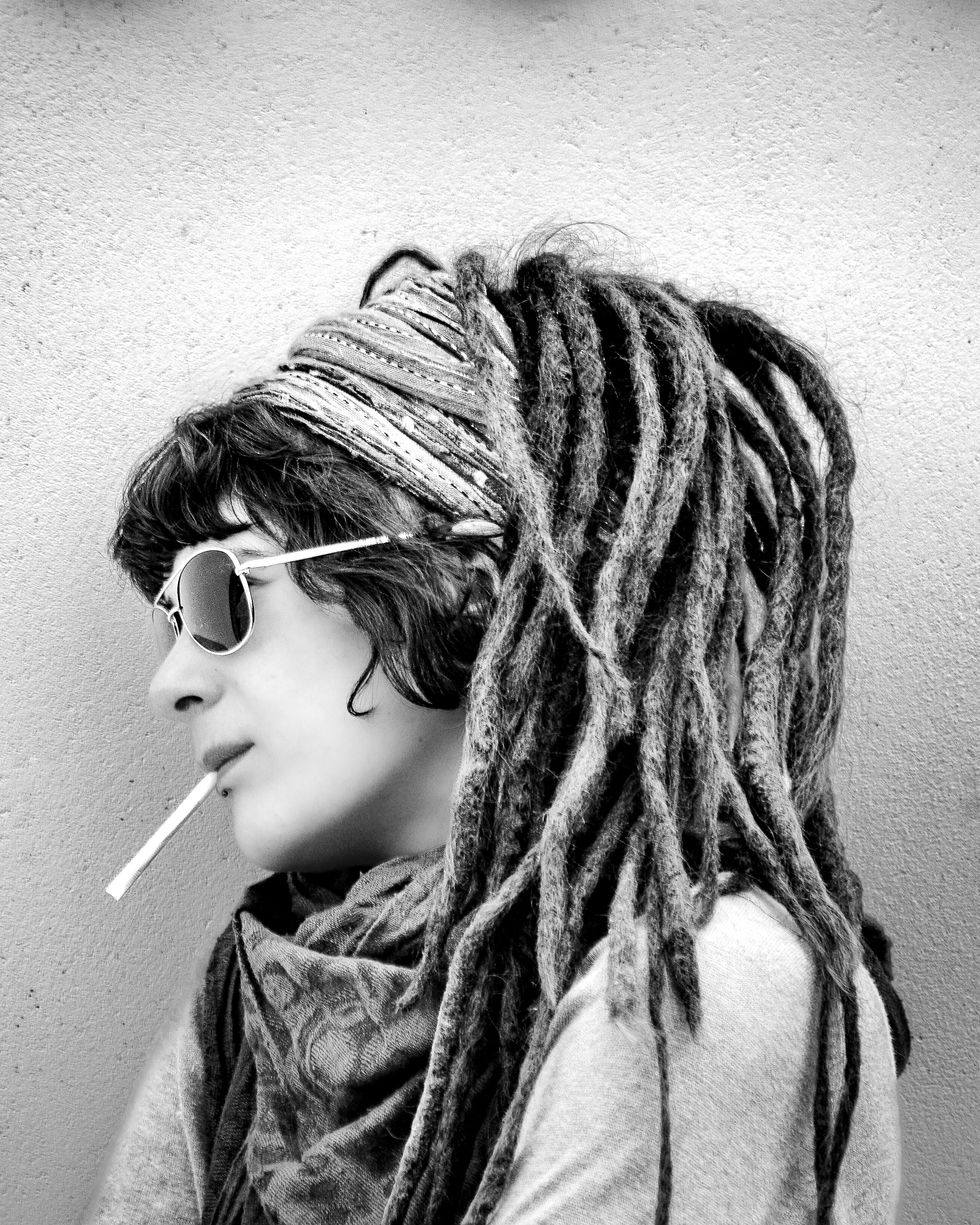 Portrait de profil en noir et blanc d'une femme portant des lunettes de soleil, un foulard et de longues dreadlocks, une cigarette à la bouche, devant un fond uni.
