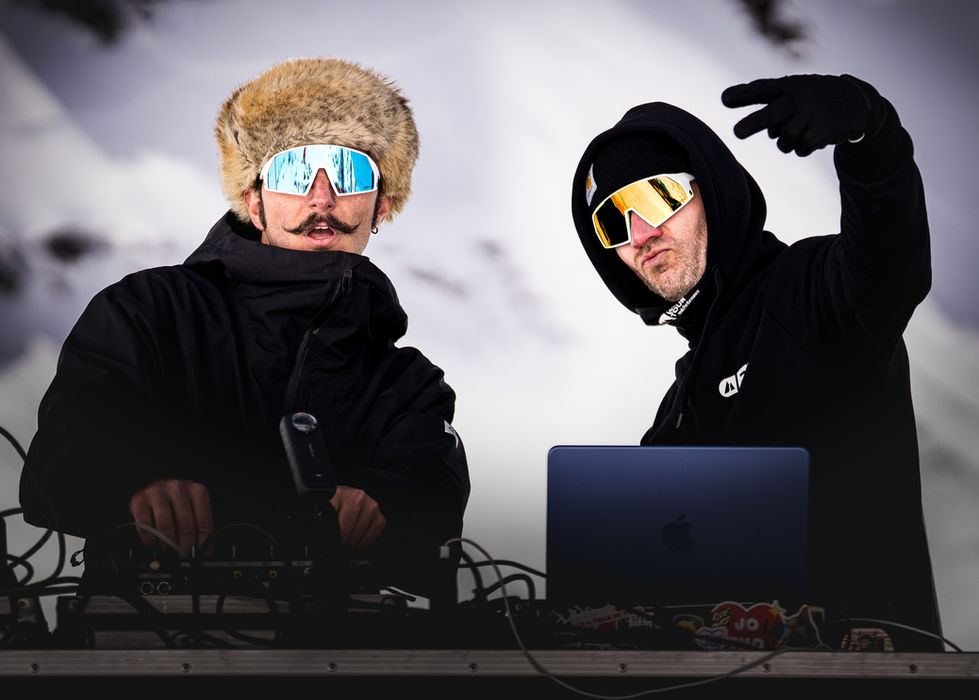 Deux DJs en tenue de ski et lunettes de soleil sur une scène enneigée. L'un porte une chapka en fourrure et une moustache guidon, tandis que l'autre, en sweat à capuche noir, fait un signe de paix derrière un ordinateur portable couvert de stickers, devant une table de mixage à Verbier en Suisse