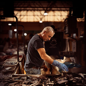 Photographie en plan moyen et en couleurs chaudes montrant un homme de profil travaillant derrière son étal dans un marché aux poissons au Portugal. Il porte un t-shirt sombre, un jean et un gant de protection bleu sur sa main droite, qu'il tend vers une multitude de poissons frais exposés au premier plan. L'arrière-plan, légèrement flou, montre l'architecture industrielle du marché avec ses lumières suspendues sous une haute structure de toit.