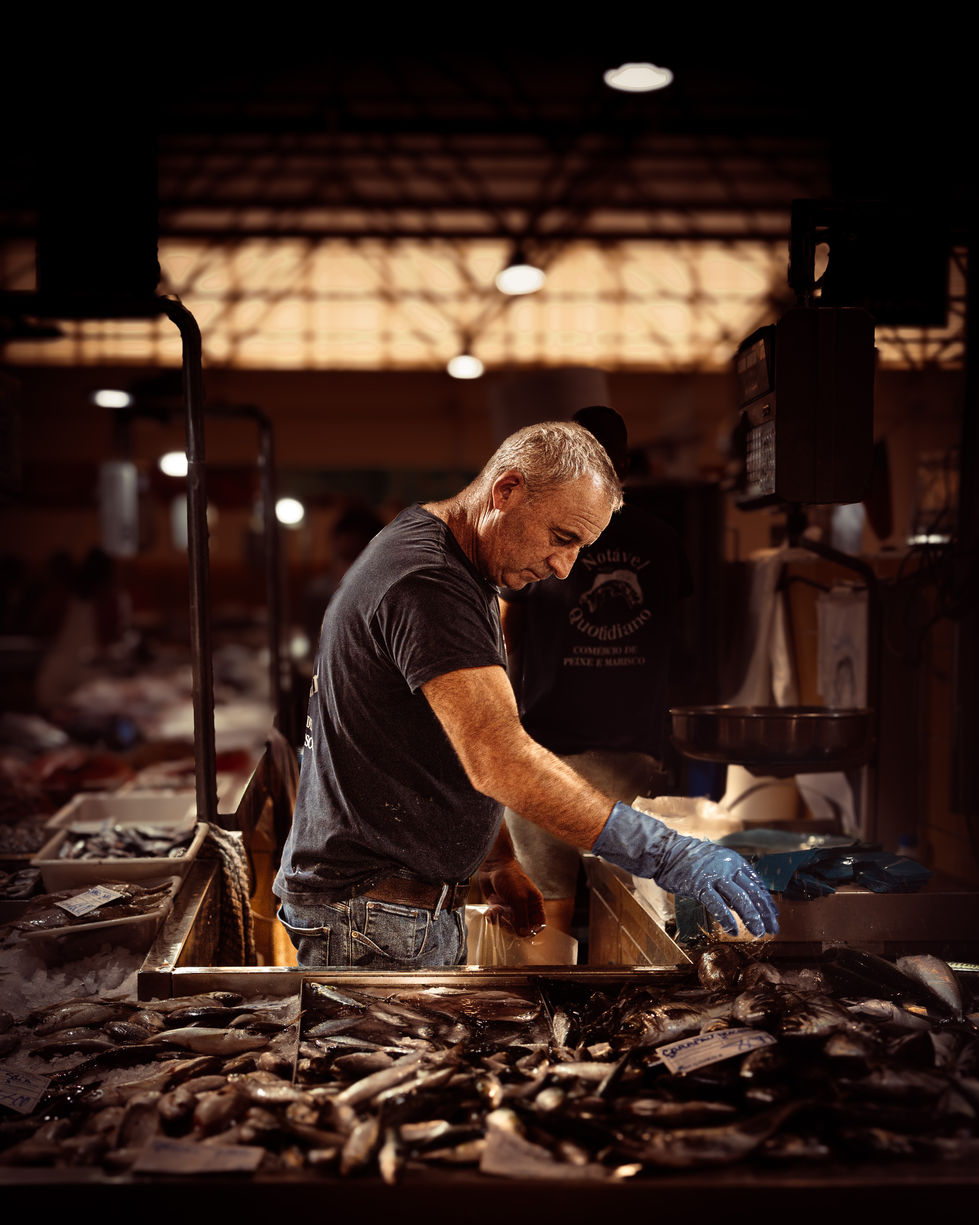 Photographie en plan moyen et en couleurs chaudes montrant un homme de profil travaillant derrière son étal dans un marché aux poissons au Portugal. Il porte un t-shirt sombre, un jean et un gant de protection bleu sur sa main droite, qu'il tend vers une multitude de poissons frais exposés au premier plan. L'arrière-plan, légèrement flou, montre l'architecture industrielle du marché avec ses lumières suspendues sous une haute structure de toit.