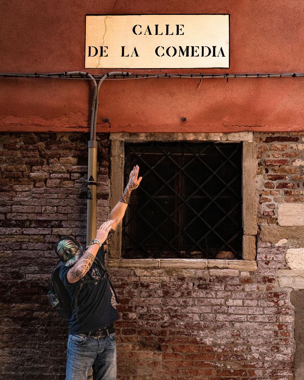 Photographie de rue en contre-plongée montrant un homme de dos portant une casquette et un sac à dos, les bras tatoués tendus dans une pose de "dab". Il se tient devant un mur en briques rouges et une fenêtre grillagée à Venise, juste en dessous d'une plaque de rue blanche où est inscrit en lettres noires "CALLE DE LA COMEDIA".
