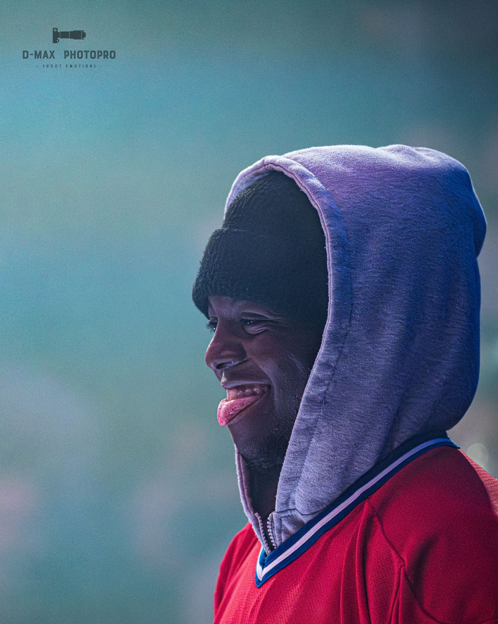 Portrait de profil rapproché d'un danseur urbain souriant et tirant la langue pendant le Red Bull Dance Your Style à Lausanne. Il porte un bonnet noir sous une capuche de sweat-shirt gris et un maillot rouge, sur un fond de scène flou aux reflets bleutés.