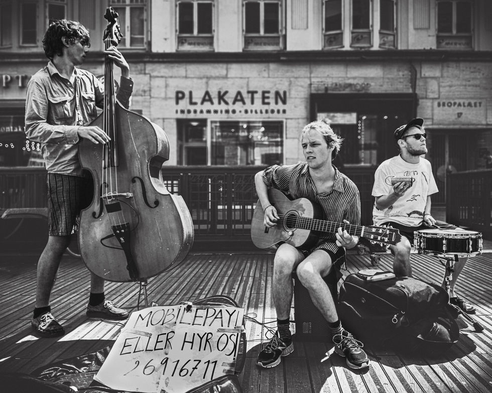 Photographie de rue en noir et blanc montrant trois jeunes musiciens jouant sur une place publique en bois à Aarhus, au Danemark. À gauche, un homme joue de la contrebasse ; au centre, un autre est assis et joue de la guitare acoustique ; à droite, un troisième bat le rythme sur une caisse claire. Au premier plan, un panneau en carton affiche un numéro de téléphone pour les paiements. En arrière-plan, la façade d'un bâtiment présente une vitrine avec l'inscription "PLAKATEN".