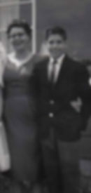 A woman and a boy in formal attire stand arm-in-arm, smiling, in front of a building with large windows. Black and white photo.