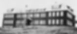 Black and white image of a large, three-story brick building with many windows, a central entrance, and a sloped roof, set against a grassy landscape.