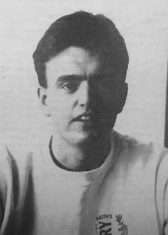 Young man in a light sweatshirt with a logo, sitting against a plain background, looking directly at the camera with a neutral expression.